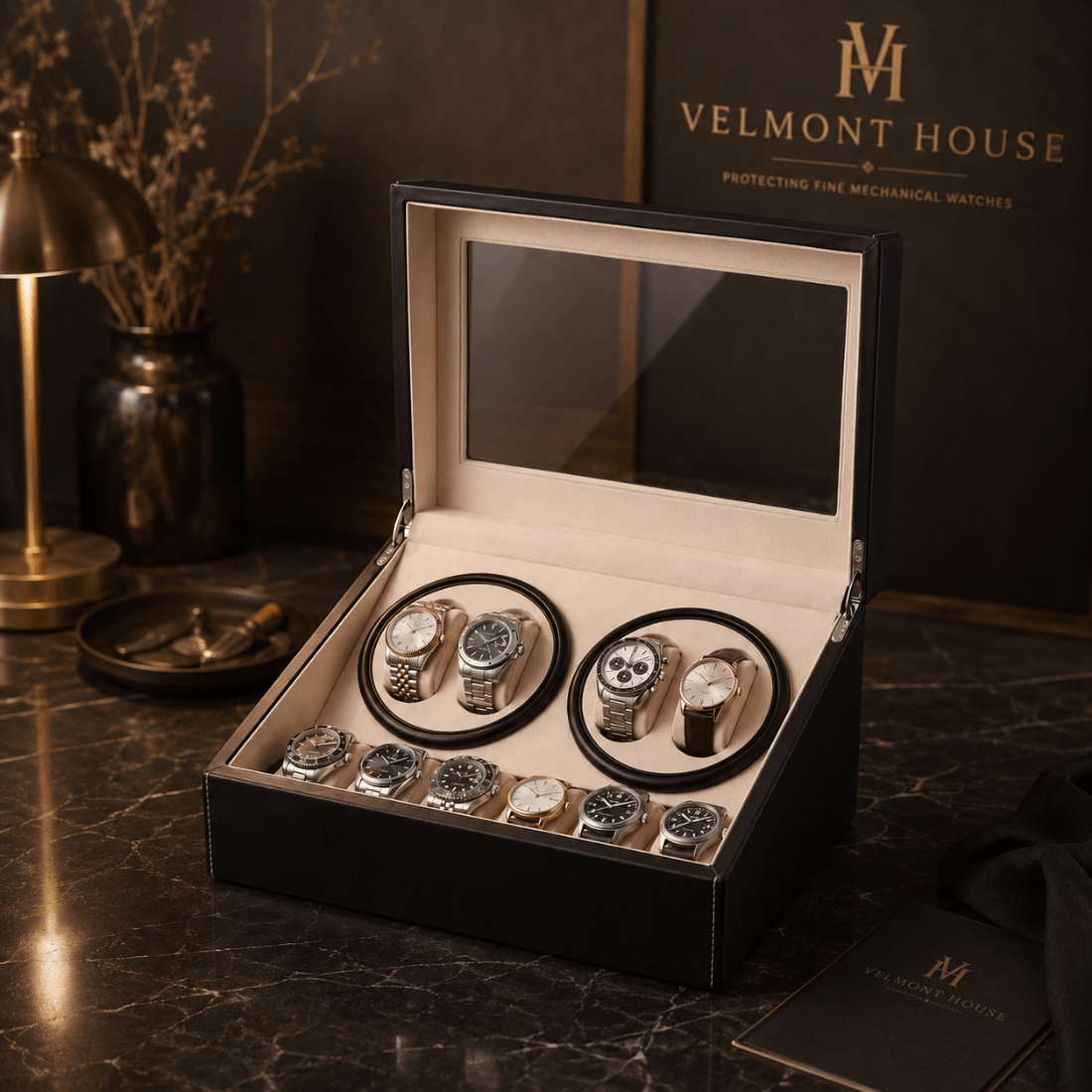 An open black velvet watch winder box displaying multiple mechanical watches, set on a dark marble surface with a branded velvet box and lamp in the background.