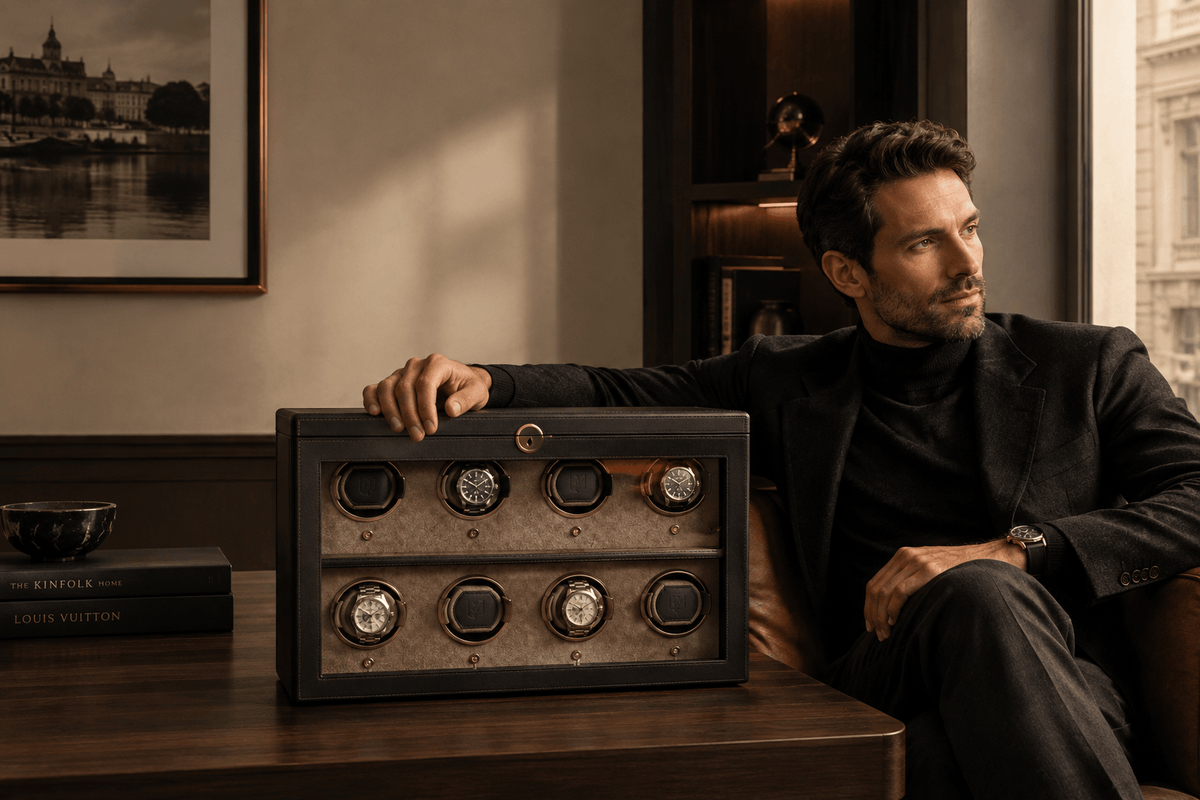 A man in a black turtleneck and blazer sits in a leather chair beside a wooden table, resting his arm on a black watch winder box with six watches visible. The room has warm neutral walls, framed artwork, and a window in the background.